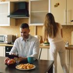 Happy young man using digital tablet computer sitting in the kitchen and talking his wife while she cooking at home in the morning